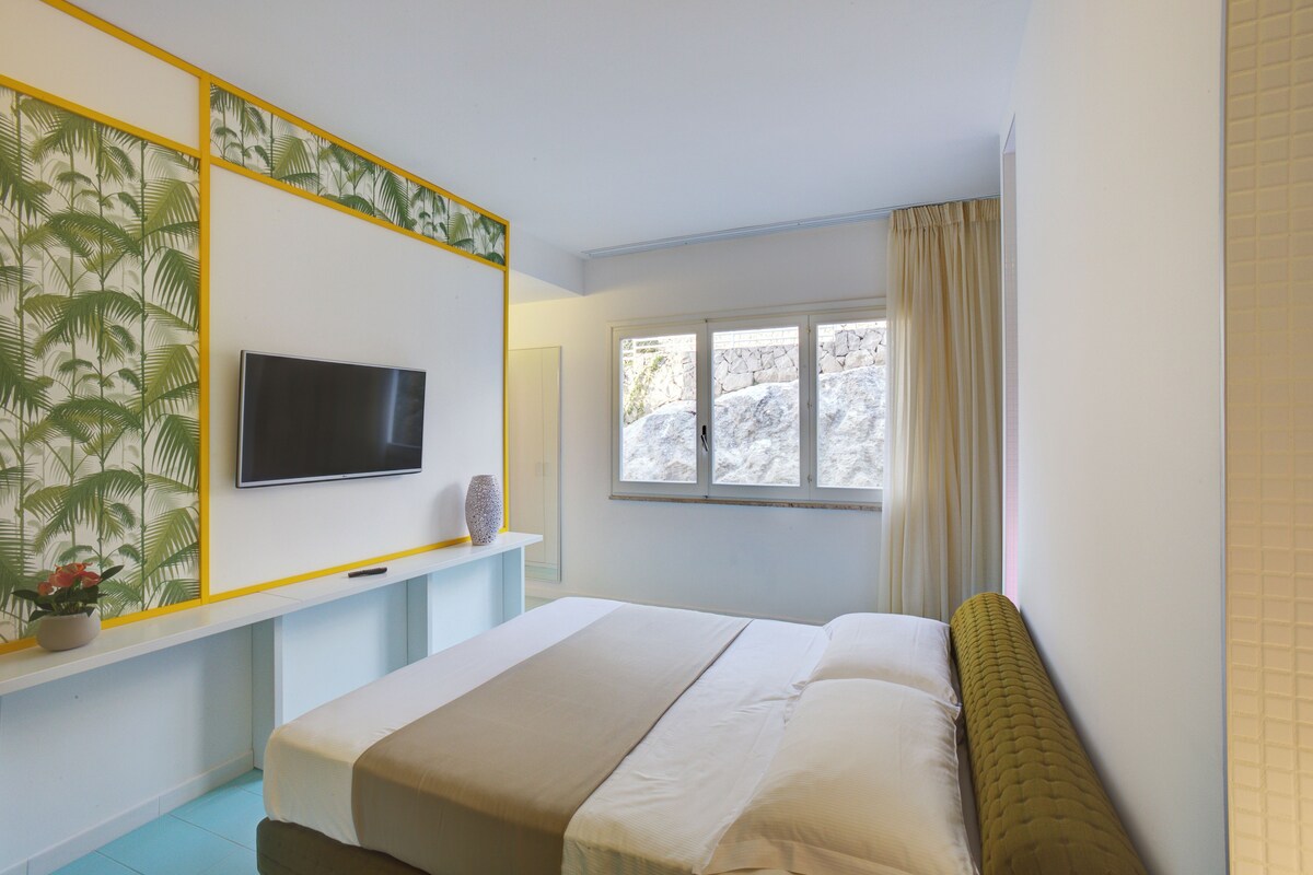 Ein Schlafzimmer mit einem Bett, einem an der Wand montierten Fernseher, einem Fenster mit Vorhängen und einem Blumentopf auf dem Nachttisch.