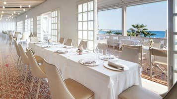 Un lungo tavolo da pranzo con tovaglie bianche e sedie, con ampie finestre che si affacciano su una spiaggia.