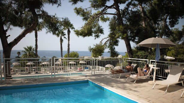 Un espace piscine doté de chaises longues et de parasols, entouré d’arbres et offrant une vue sur la mer.