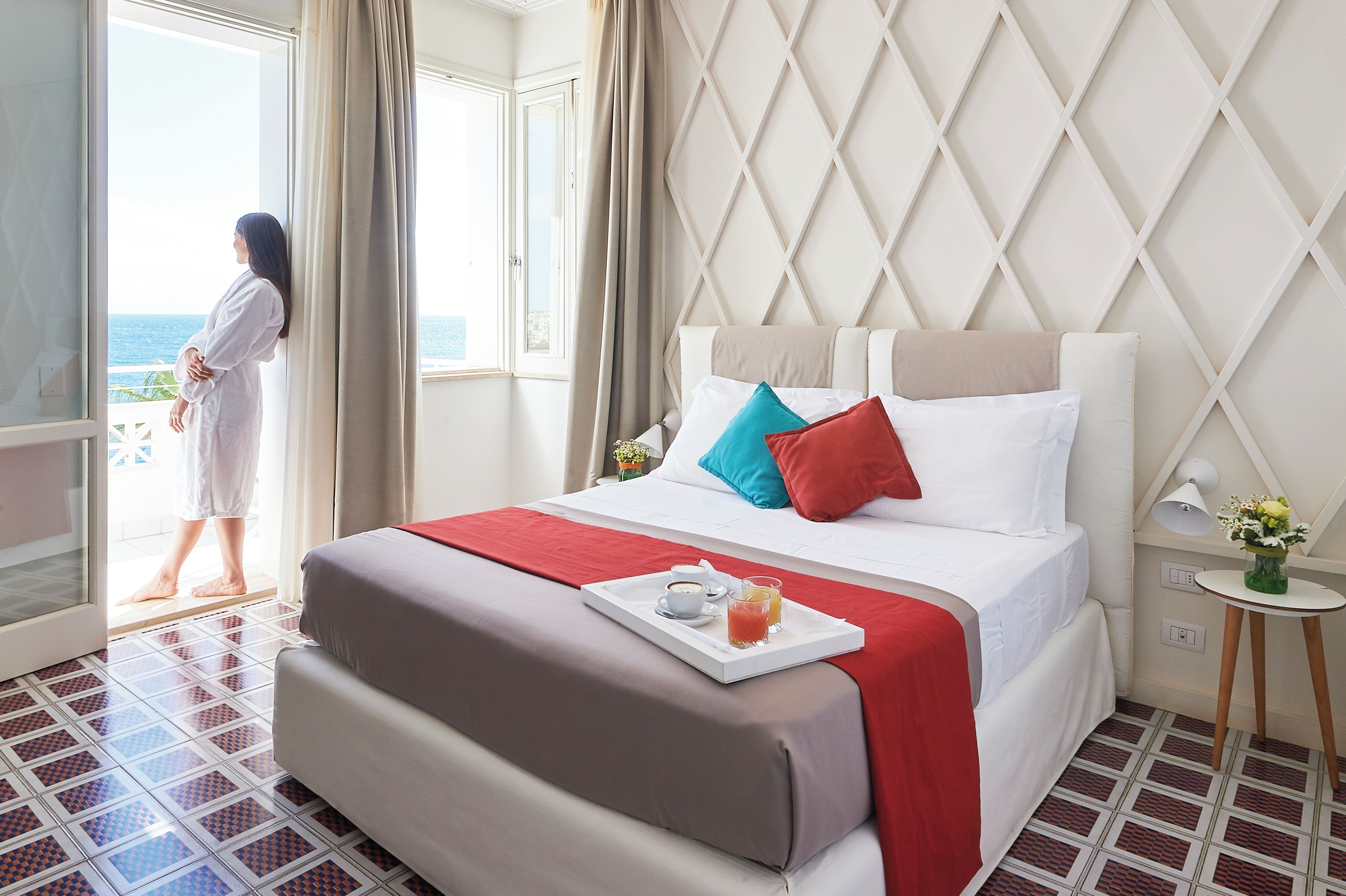A hotel room with a bed, a tray with breakfast, a window with a view, and a person standing by the window.