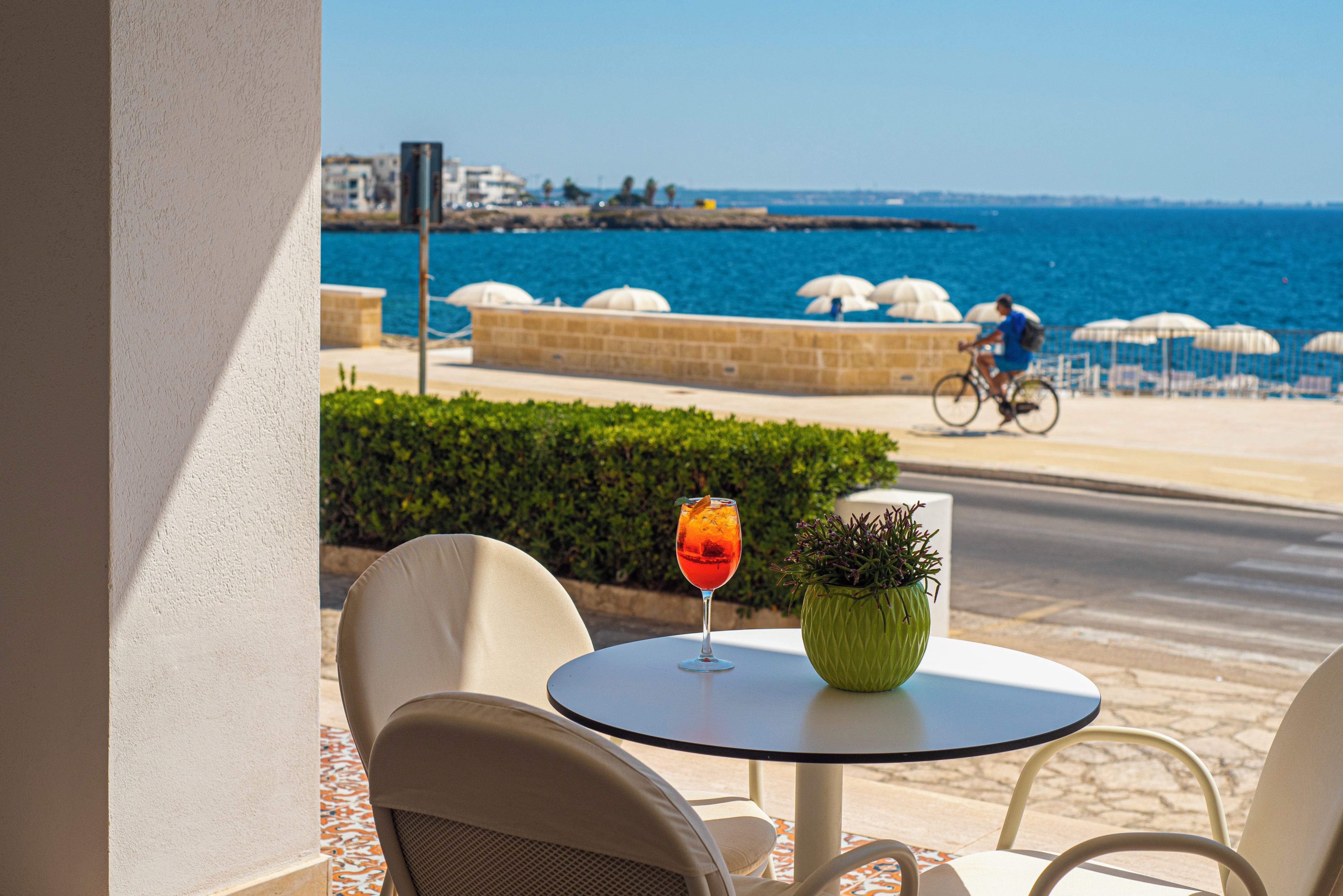 A table with a drink and a plant, overlooking a coastal area with a cyclist in the background.