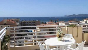 Un balcon avec une table et des chaises blanches, donnant sur une zone côtière avec des bâtiments et la mer.