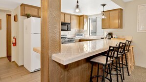A modern kitchen with wooden cabinets, a white refrigerator, a microwave, a stove, and a large island with bar stools. - Summit Ski and Bike Trails #2574 (Big Bear Lake)