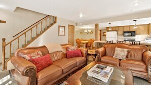 A spacious living room with a brown leather sofa, a coffee table, and a dining area with a wooden table and chairs. A staircase with wooden railings is visible in the background. - Summit Ski and Bike Trails #2574 (Big Bear Lake)