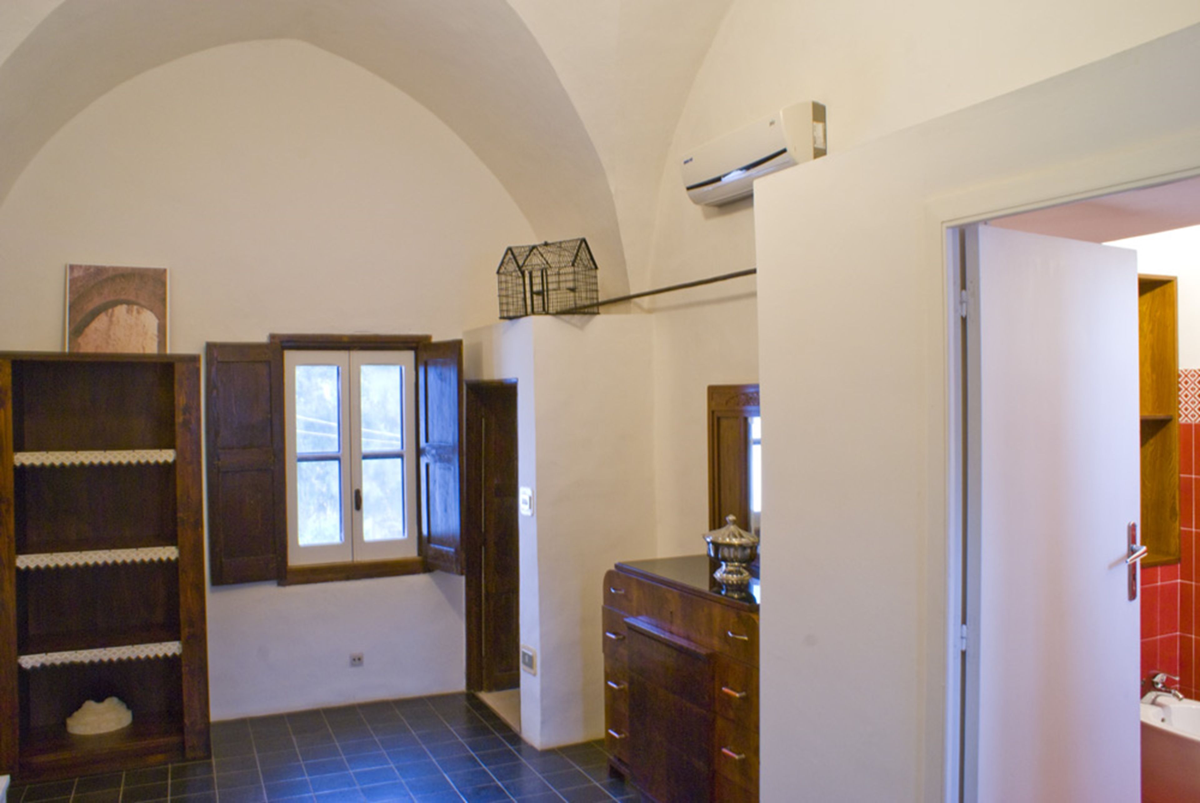 A room with a tiled floor, a wooden wardrobe, a small window with a view, and an air conditioning unit.