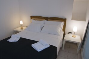 Een net opgemaakt bed met witte lakens en twee opgevouwen handdoeken op een zwart dekbed. Er zijn twee nachtkastjes, elk met een lamp.