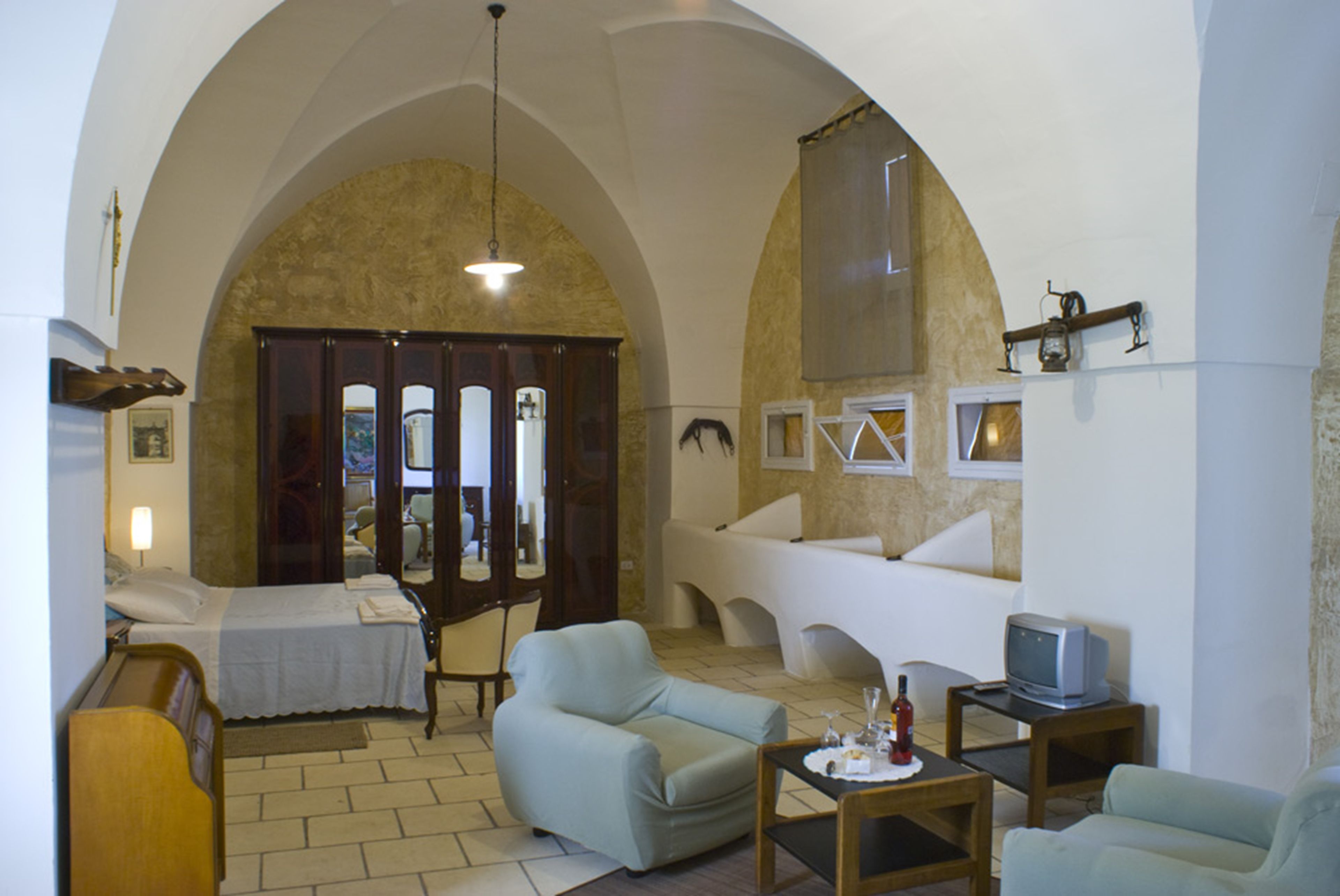 A living room with a bed, two armchairs, a small table, a television, and a large arched doorway.