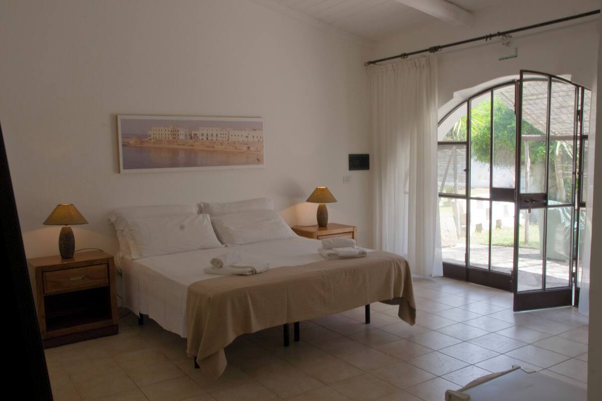 Habitación de hotel con cama, dos mesitas de noche con lámparas, un cuadro en la pared y un ventanal con cortinas.