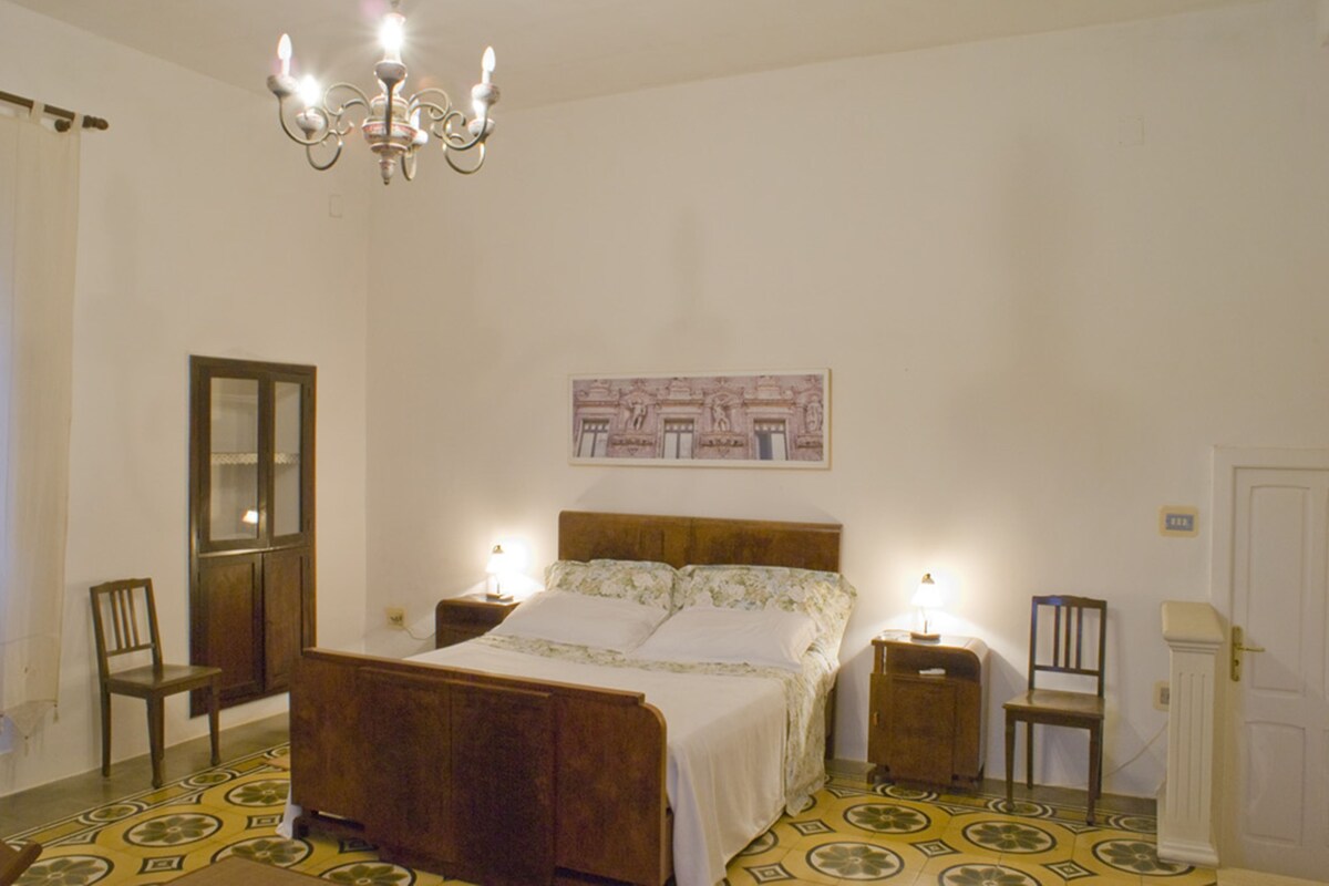 Een slaapkamer met een houten bed, twee nachtkastjes, een kroonluchter, een spiegel en een deur.