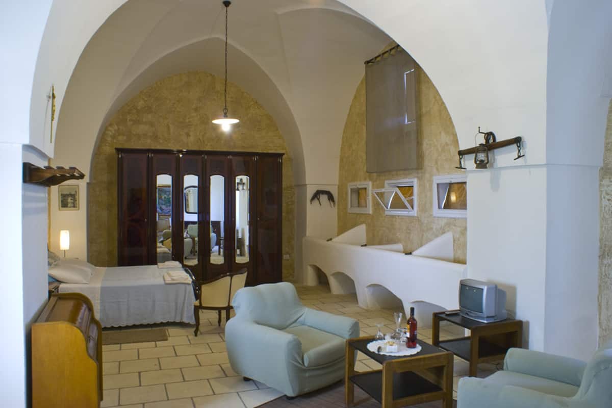 Una sala de estar con una cama, dos sillones, una mesita, un televisor y un amplio arco de entrada.
