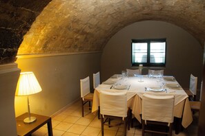 Un comedor con techo abovedado de piedra, una mesa dispuesta para seis personas con manteles blancos y sillas, y una lámpara sobre una mesita auxiliar.