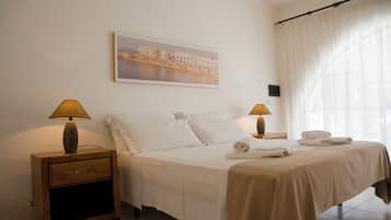A neatly made bed with white linens, two bedside lamps, a nightstand with a drawer, a framed picture on the wall, and a window with sheer curtains.