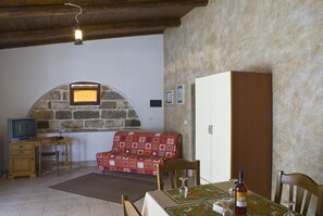 A cozy living area with a stone wall, a wooden ceiling, a television, a sofa, a cabinet, and a dining table with a bottle and glasses.