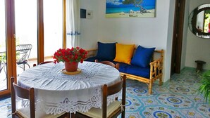 Una habitación con una mesa redonda cubierta por un mantel de encaje, una planta en maceta con flores rojas, un sofá azul con cojines amarillos y un cuadro con una escena de playa en la pared.