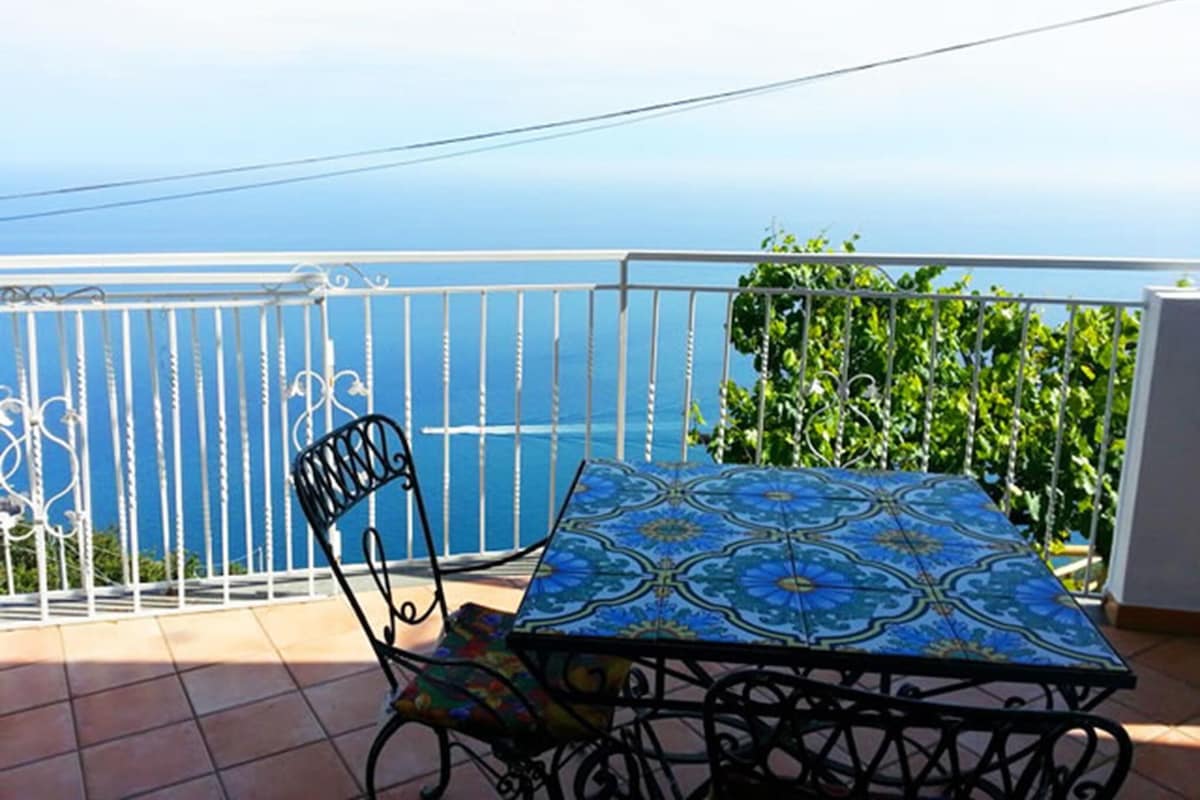 A balcony with a tiled table and chairs, a metal railing, and a view of the sea.