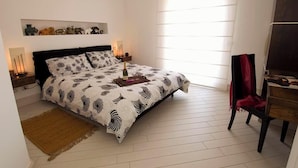 Ein Schlafzimmer mit einem Bett, Nachttischen, einem Hocker, einem Sessel, einem Regal mit Dekoration und einem Fenster.