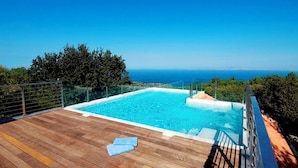 Ein rechteckiger Pool mit Holzdeck und Metallgeländer, umgeben von Grünpflanzen und mit Blick auf ein Gewässer.