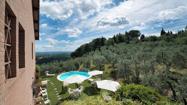 A pool with white umbrellas, a brick building, and a hillside with trees. - Casa Lari (San Gimignano)