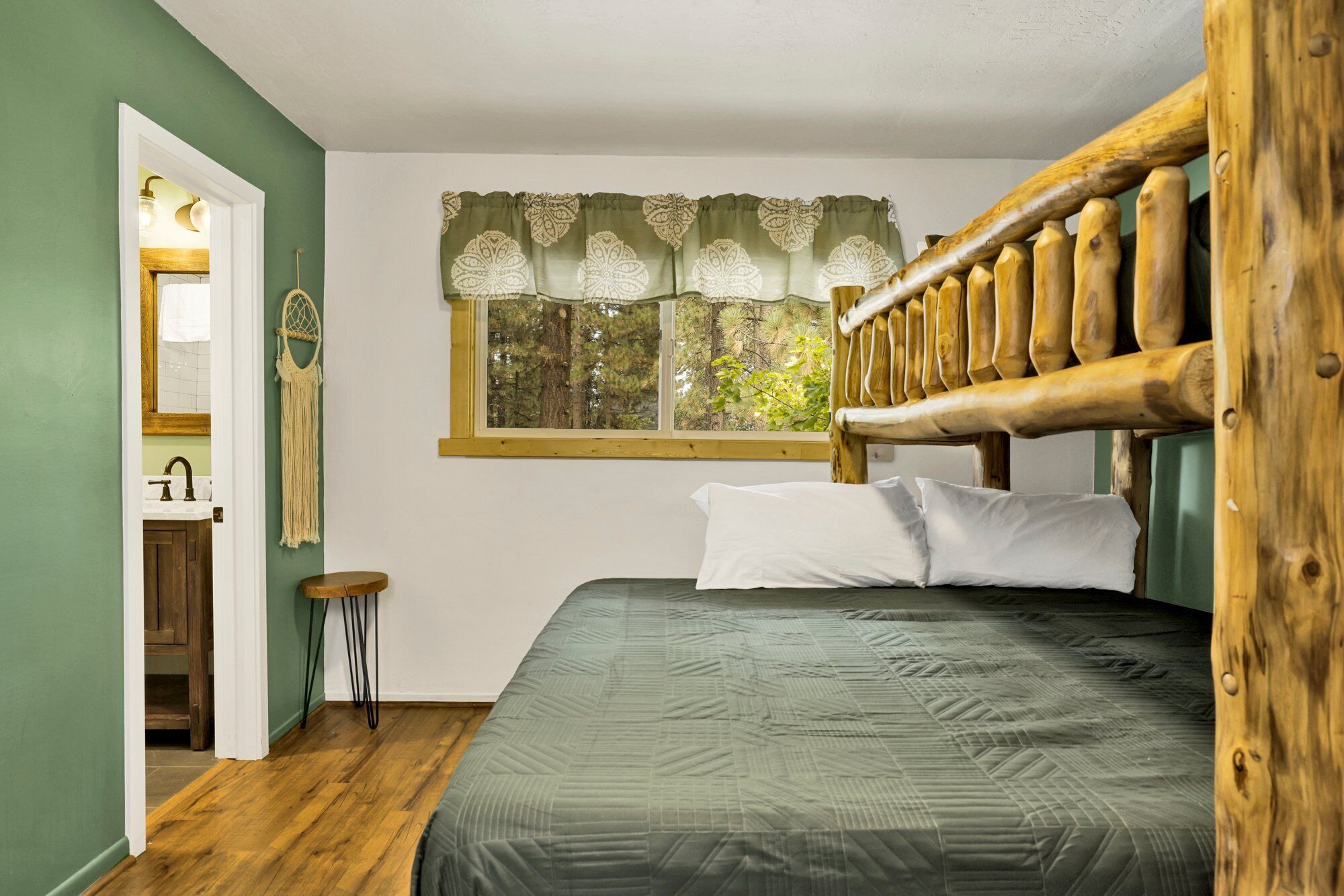 A bedroom with a wooden headboard, a bed with a green quilt, a window with a patterned curtain, a wooden stool, and a mirror with a wall hanging.