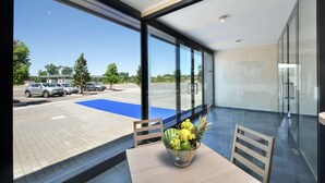 Une chambre d’hôtel moderne avec de grandes fenêtres, donnant sur un parking, et une table sur laquelle se trouve un panier de fruits.