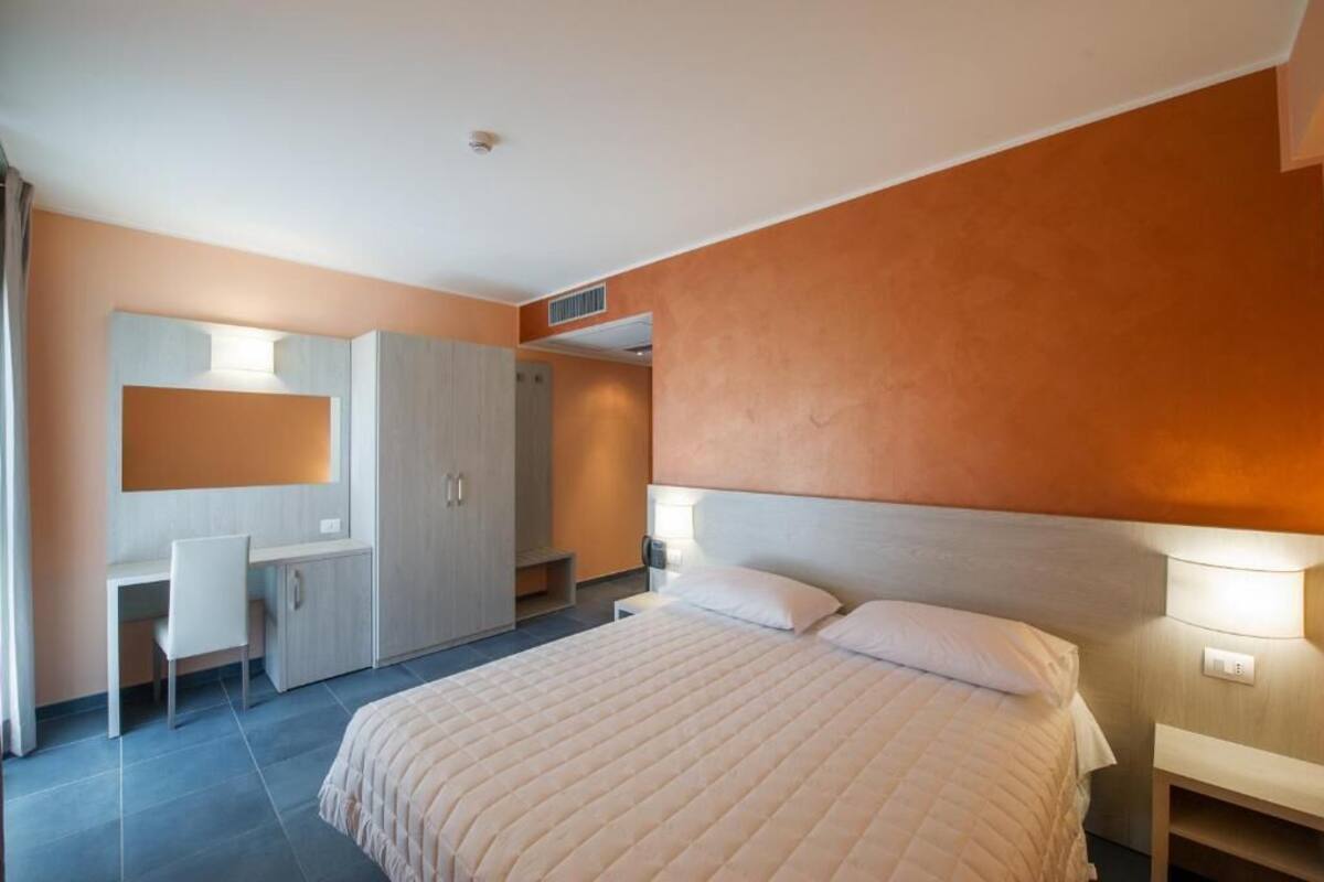 Quarto com cama, mesa de cabeceira, luminária, guarda-roupa, escrivaninha, cadeira e espelho.