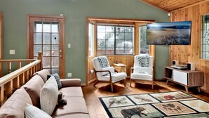 Un salon chaleureux avec un plafond en bois, une grande fenêtre, un canapé, deux fauteuils, un téléviseur et un tapis.