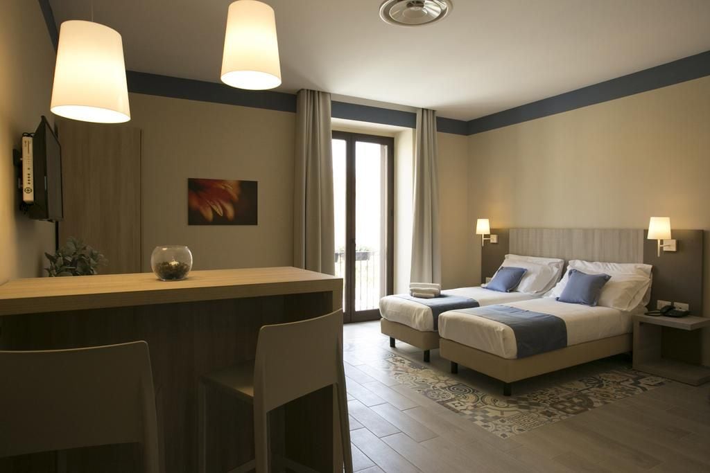 A hotel room with a bed, two bedside tables, a desk with a chair, a TV, and a large window with curtains.