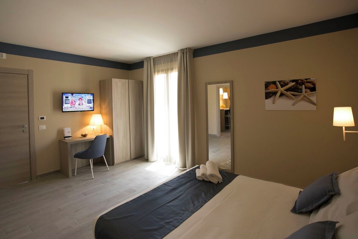 Quarto de hotel com cama, escrivaninha, cadeira, televisão e espelho.