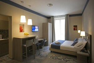 A hotel room with a bed, desk, chairs, a television, and a window with curtains.