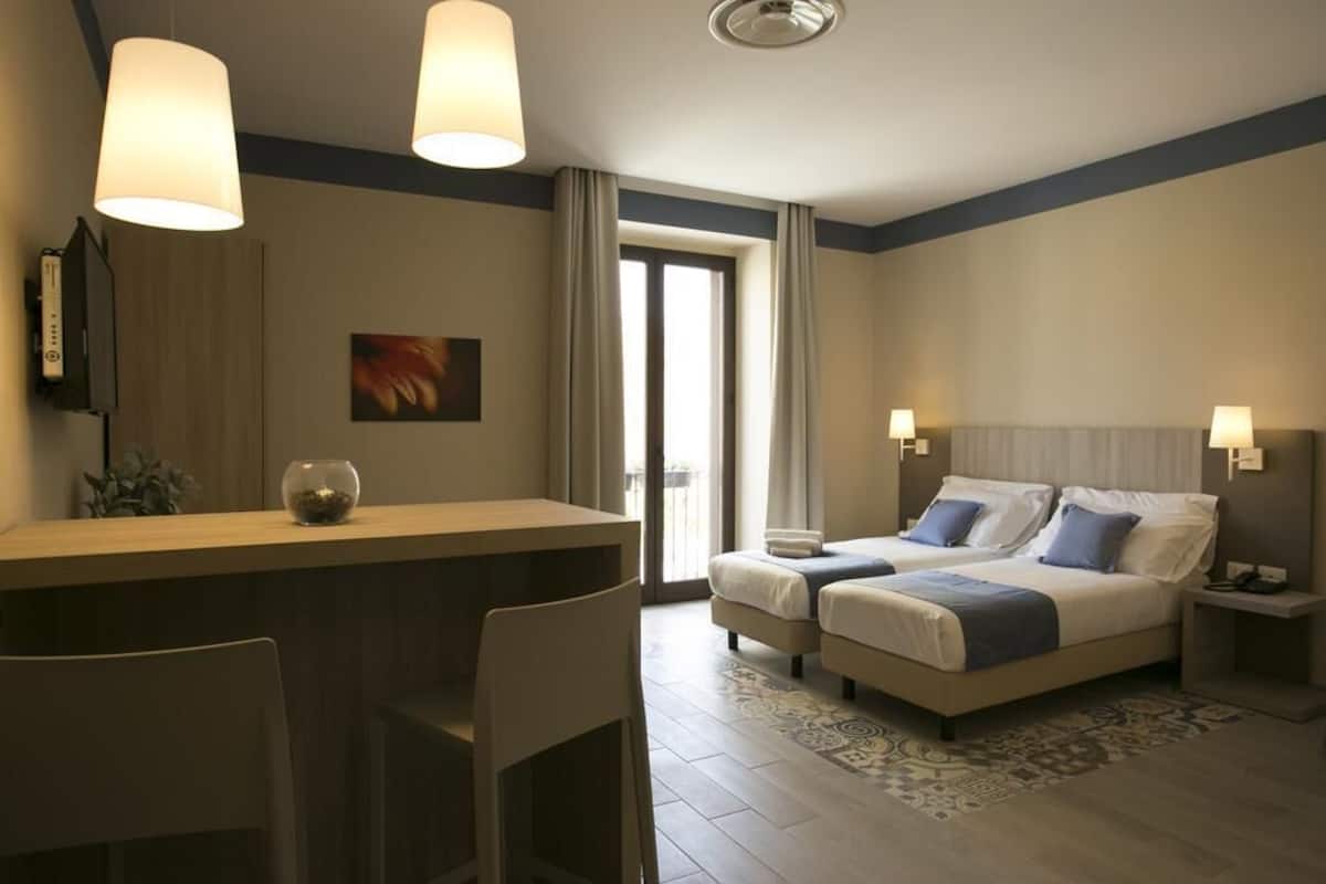 Habitación de hotel con cama, dos mesitas de noche, un escritorio con silla, televisor y una ventana grande con cortinas.