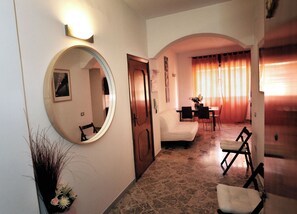 A hallway with a round mirror, a white sofa, a dining table with chairs, and a window with curtains. - Le Case Della Costa (Maiori)