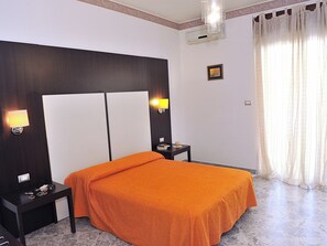 A hotel room with a bed, two bedside tables, a nightstand, a wall-mounted air conditioner, a framed picture, and a window with sheer curtains. - Le Case Della Costa (Maiori)
