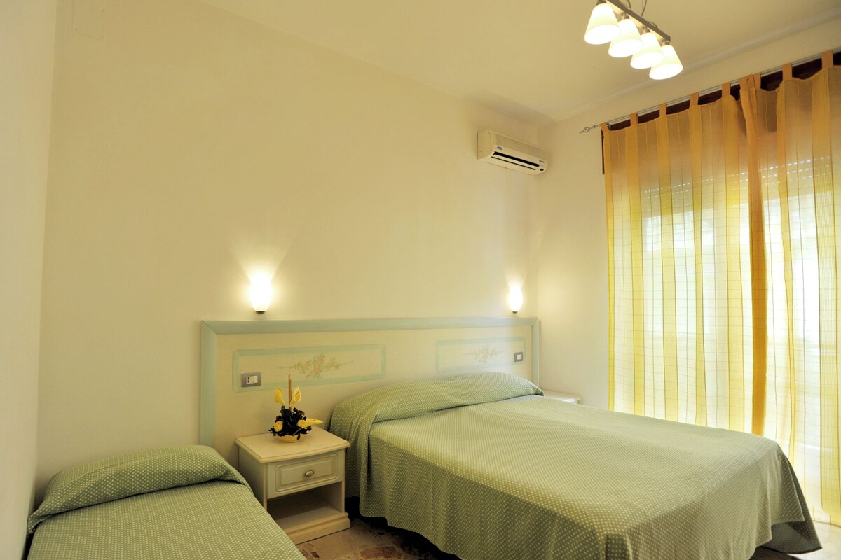 A room with two beds, a nightstand with a lamp, an air conditioning unit, and a window with curtains.