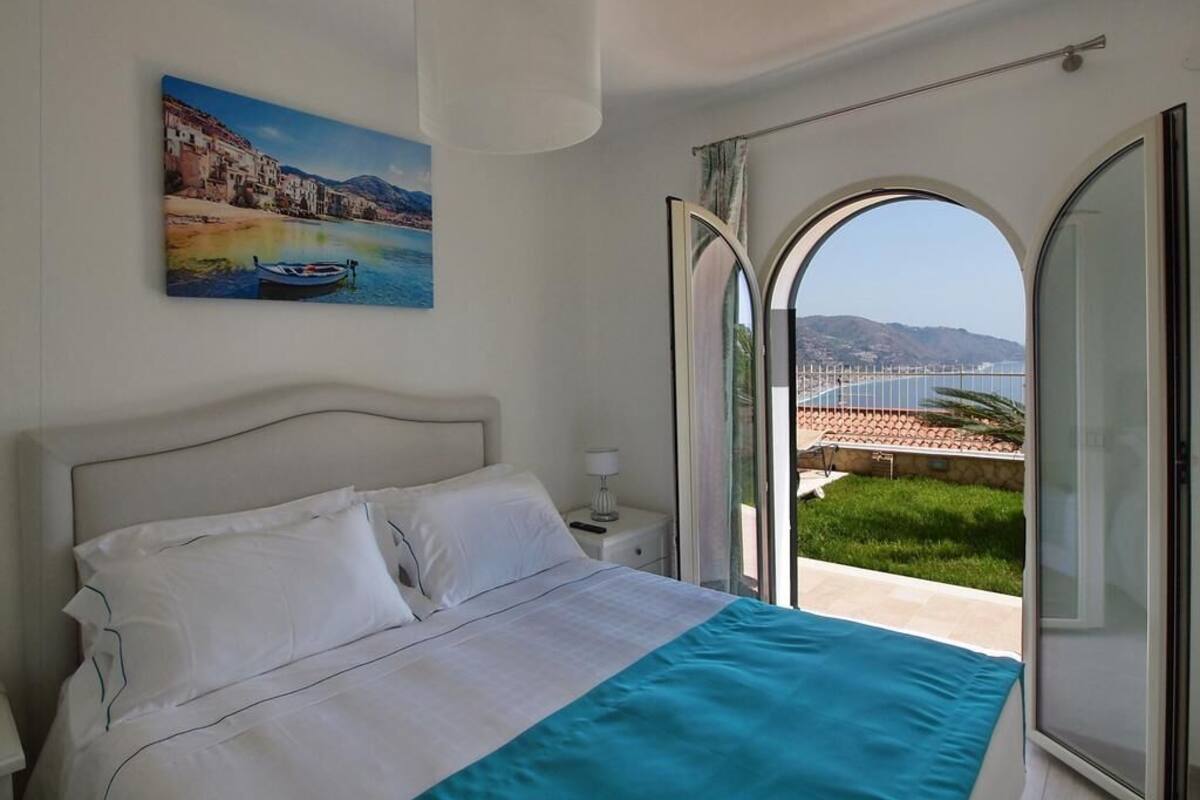 A bedroom with a bed, a nightstand, a painting, and a view through an open door.