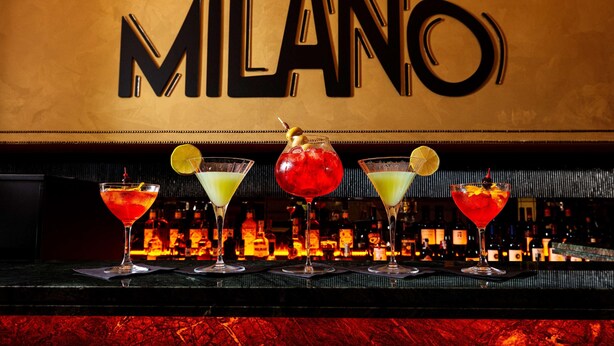 Een bar met een 'MILANO' bord, diverse cocktails en een assortiment drankflessen.