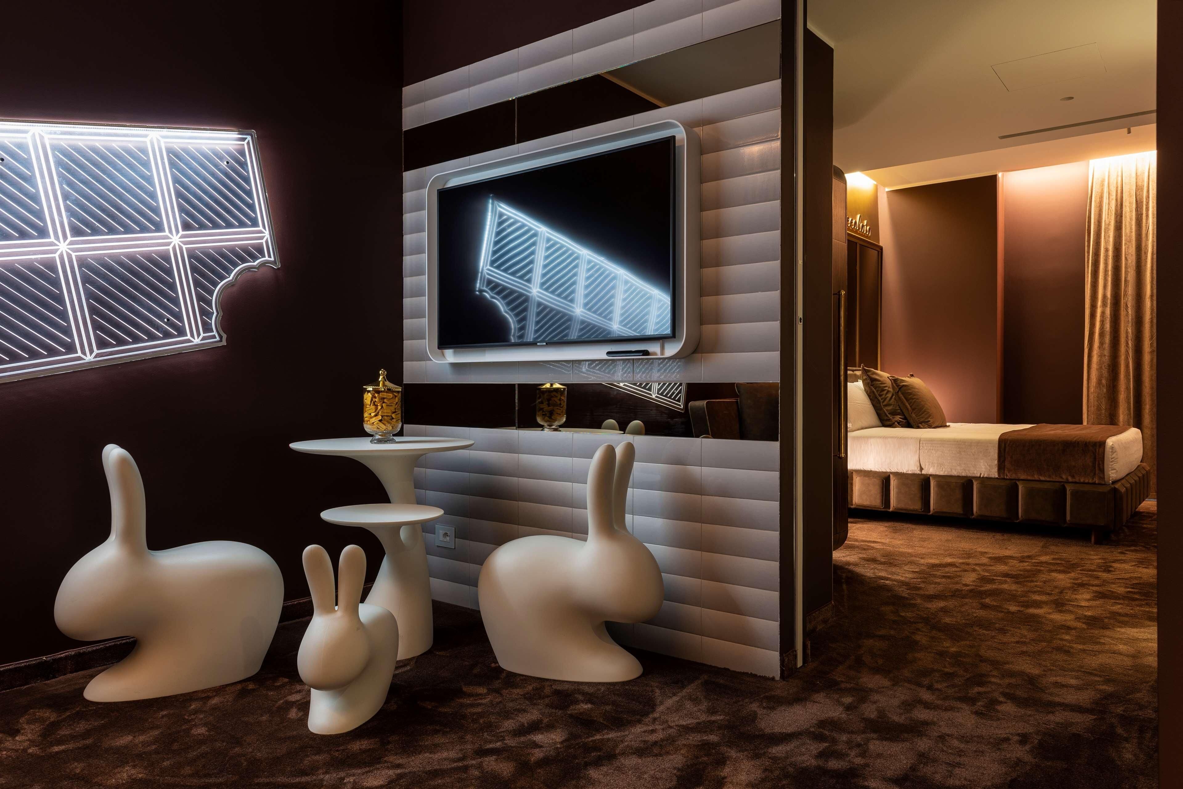Een moderne kamer met een grote flatscreen televisie aan de muur, een klein rond tafeltje, twee witte stoelen in de vorm van konijnen en een bed met een bruin hoofdeinde.