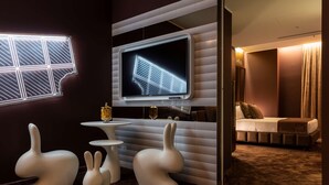 A modern room with a large flat-screen TV mounted on the wall, a small round table, two white bunny-shaped chairs, and a bed with a brown headboard. - Odsweet Duomo Milano Hotel (Milan)
