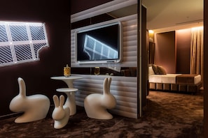 Une chambre moderne dotée d’un grand téléviseur  à écran plat fixé au mur, d’une petite table ronde, de deux chaises blanches en forme de lapin et d’un lit avec une tête de lit marron.