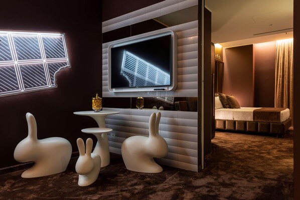 Una habitación moderna con una gran televisión de pantalla plana montada en la pared, una pequeña mesa redonda, dos sillas blancas con forma de conejo y una cama con cabecera marrón.
