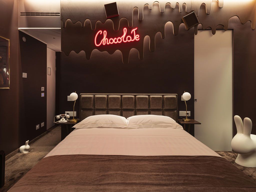 Habitació d'hotel amb decoració inspirada en el xocolata, que inclou un cartell de neó, un llit mull i càlid, làmpades laterals i una estàtua de conill blanc.