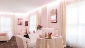 Een hotelkamer in roze kleurstelling, met een bank, een klein tafeltje met een bloevas en een bed met een roze deken.