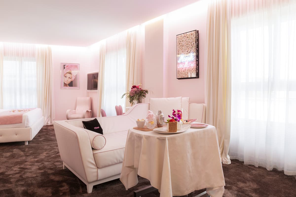 Une pièce décorée dans des tons roses, avec un lit, deux canapés, une table ornée d'un vase de fleurs et un panneau lumineux au néon.