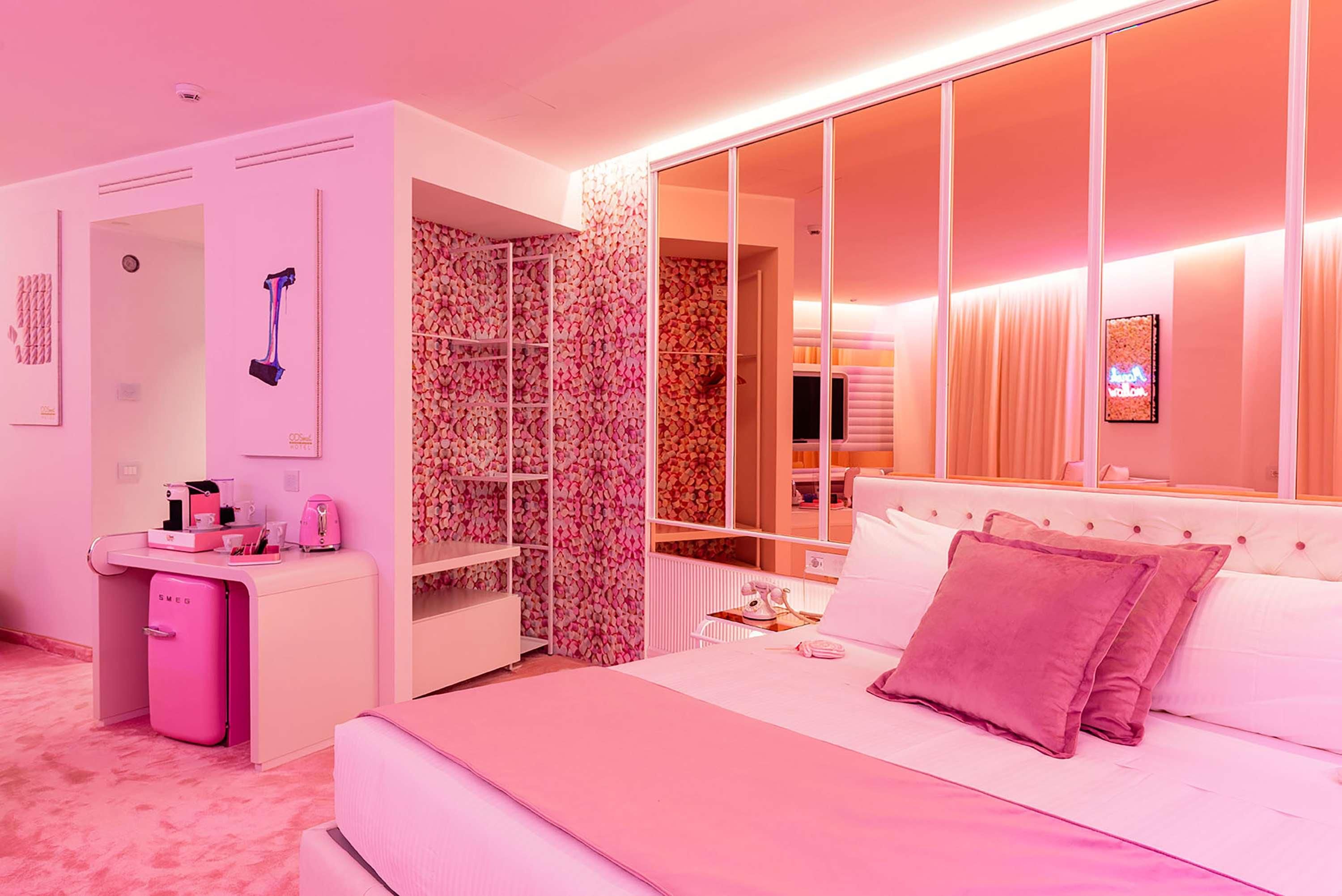 Ein Hotelzimmer in Rosa mit Bett, Schminktisch mit Spiegel, Kleiderschrank und großem Fenster.