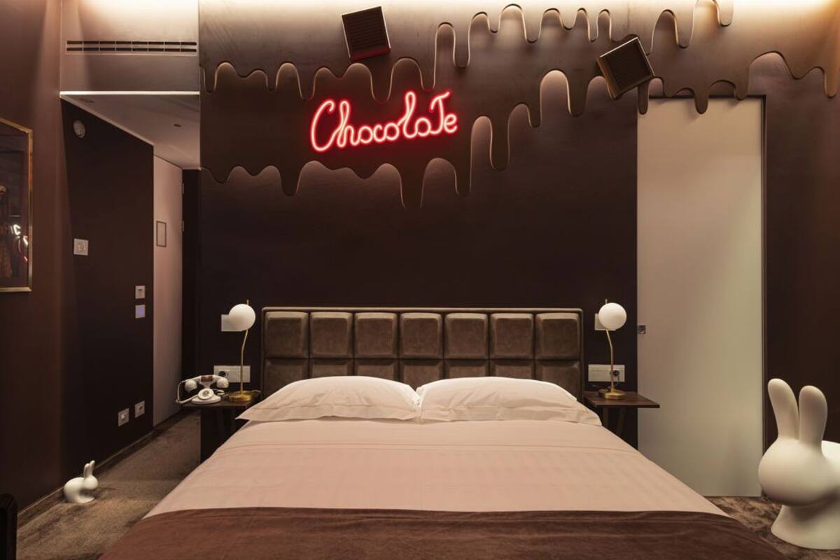 Une chambre d'hôtel décorée sur le thème du chocolat, avec notamment un néon, un lit moelleux, des lampes de chevet et une statue de lapin blanc.
