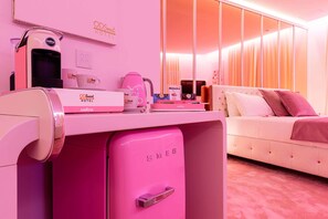 Een hotelkamer in roze tinten, met een bed, een bureau met een koffieapparaat en een minibar.