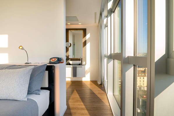 Une chambre d’hôtel moderne, dotée d’un lit, d’une table de chevet avec une lampe, d’une vue sur la ville grâce à de grandes fenêtres, et d’une salle de bain avec un lavabo et un miroir.