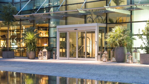Una entrada de hotel moderna con puertas de vidrio, plantas en macetas y una piscina reflectante.