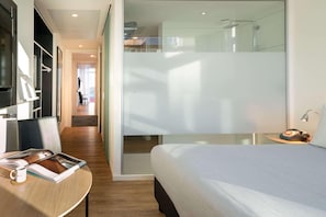 Una habitación de hotel moderna con cama, un escritorio con una revista, una taza de café y una mesita de noche con una lámpara.