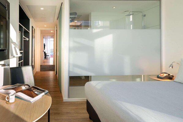 Ein modernes Hotelzimmer mit einem Bett, einem Schreibtisch mit einer Zeitschrift, einer Tasse Kaffee und einem kleinen Tisch mit einer Lampe.