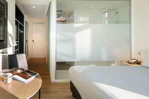 Een moderne hotelkamer met een bed, een bureau met een boek en een mok, een klein tafeltje met een lamp en een glazen scheidingswand.